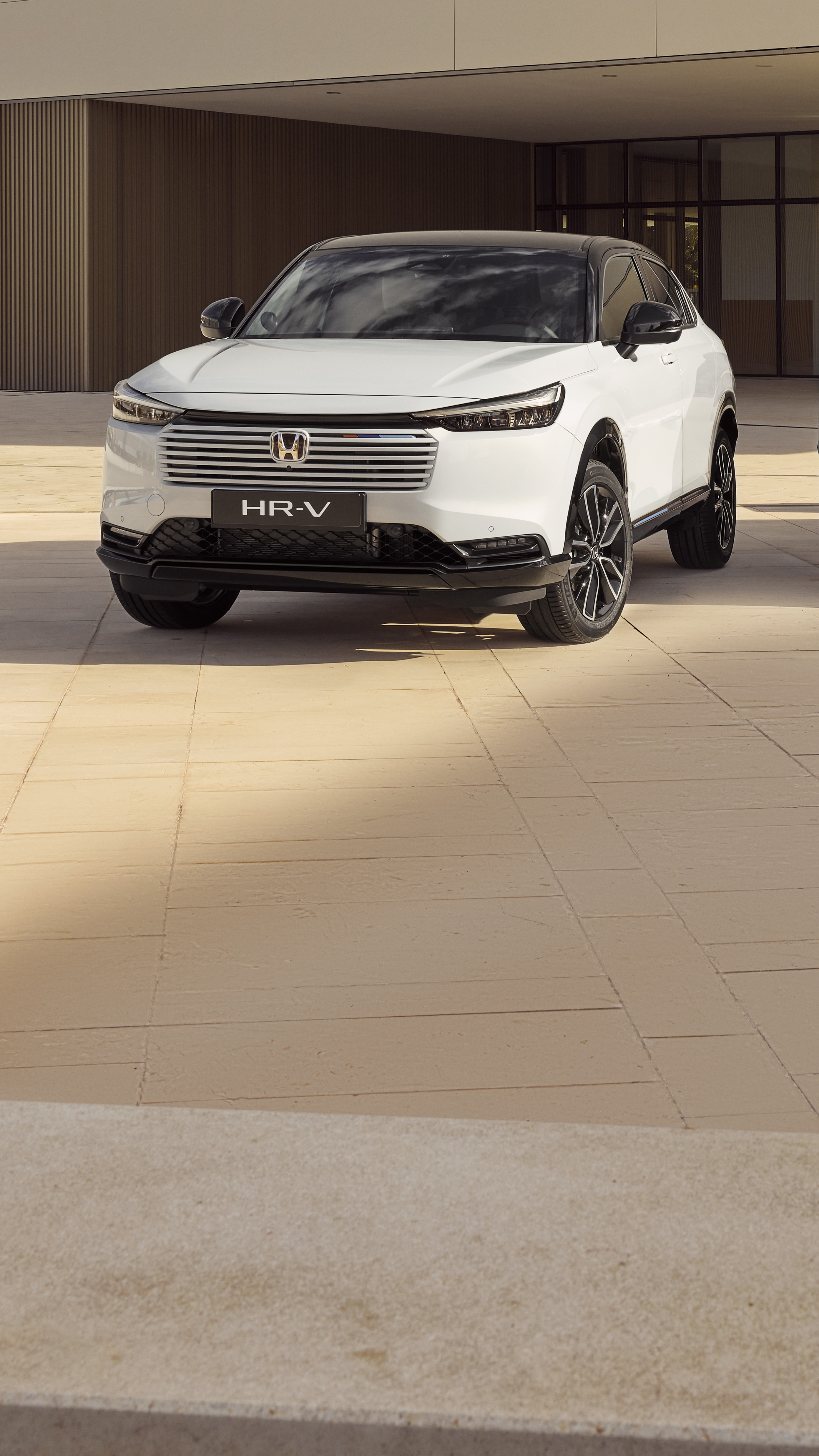 Een witte Honda HR-V SUV geparkeerd voor een modern gebouw met beige betegelde muren. Het voertuig staat schuin gepositioneerd, wat het strakke ontwerp en de karakteristieke grille benadrukt.