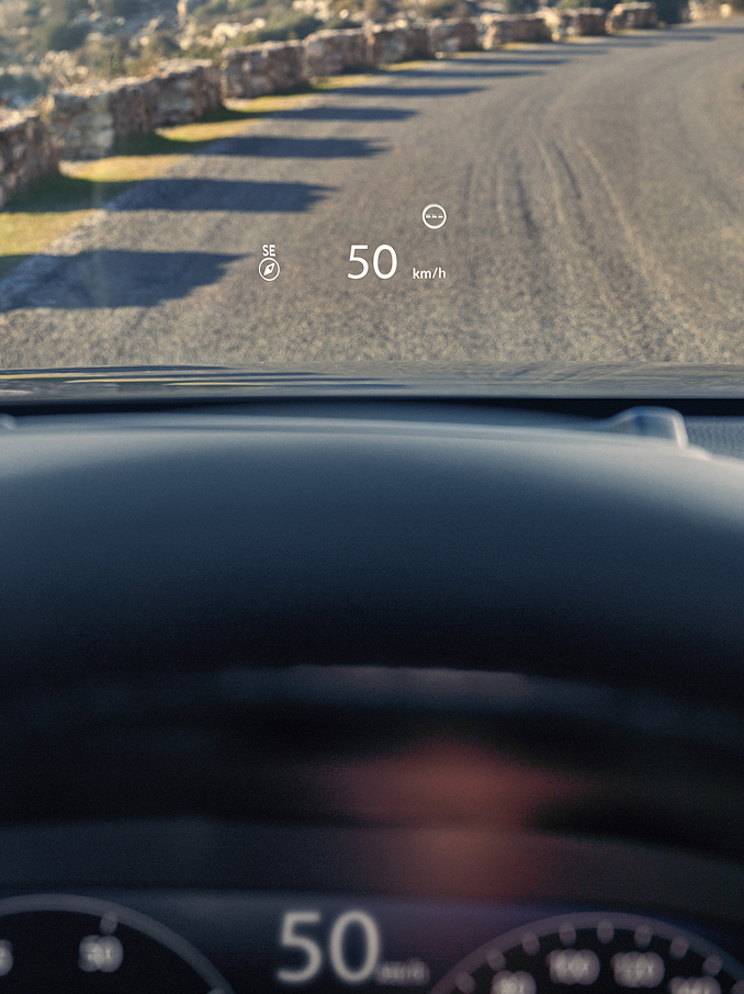 Een eerste-persoonszicht vanuit de bestuurdersstoel toont een Head-Up Display (HUD) die "50 km/h" en een kompas op de voorruit projecteert boven een grindweg.