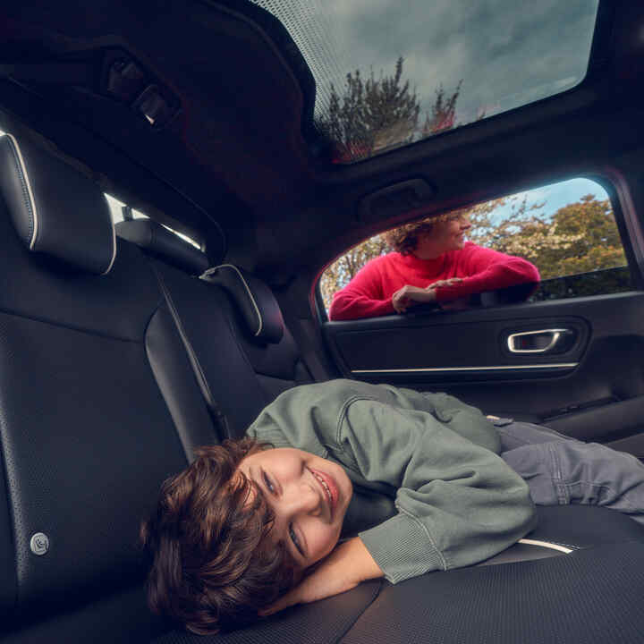 Kind liggend op de achterzetel terwijl moeder voor het raam staat van Honda e:Ny1.