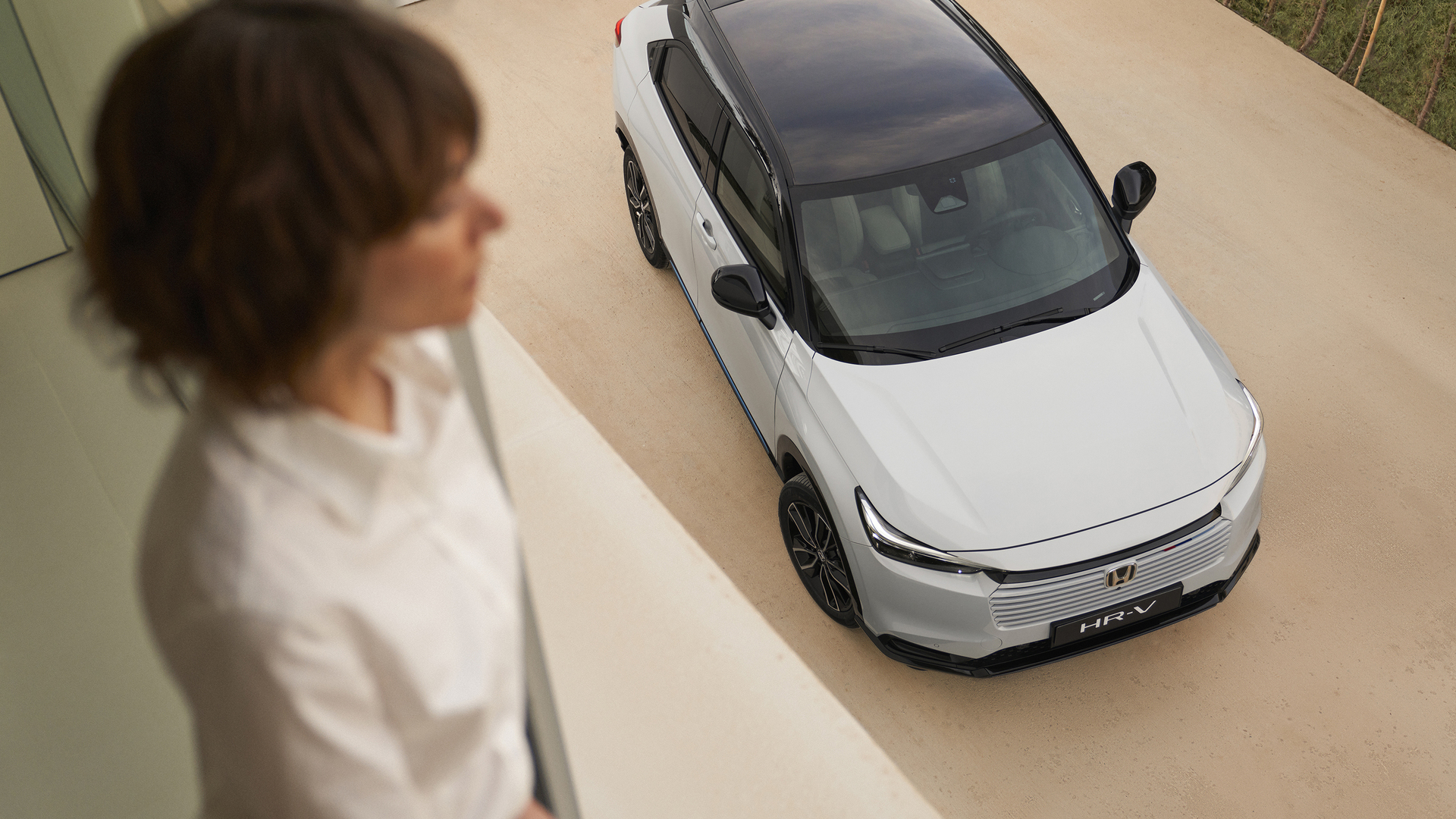 An overhead view of a white Honda HR-V with a black roof parked on a light-colored driveway, while a blurred person stands indoors in the foreground looking down toward the car.