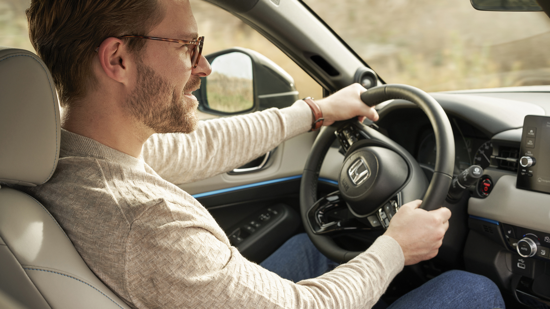 A man wearing glasses drives a Honda HR-V, shown from the front passenger seat, with both hands on the steering wheel and the dashboard visible.