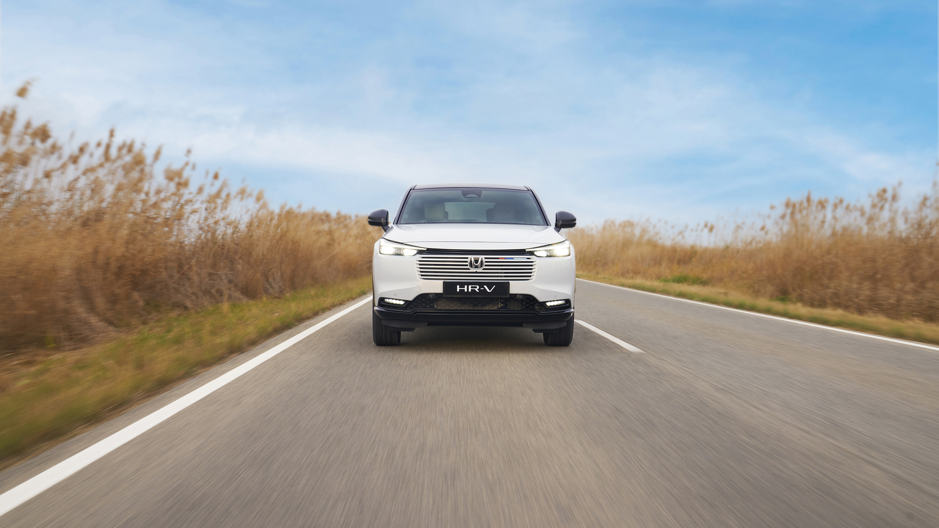 A white Honda HR-V drives toward the camera on a two-lane road, surrounded by tall golden grass under a blue sky.