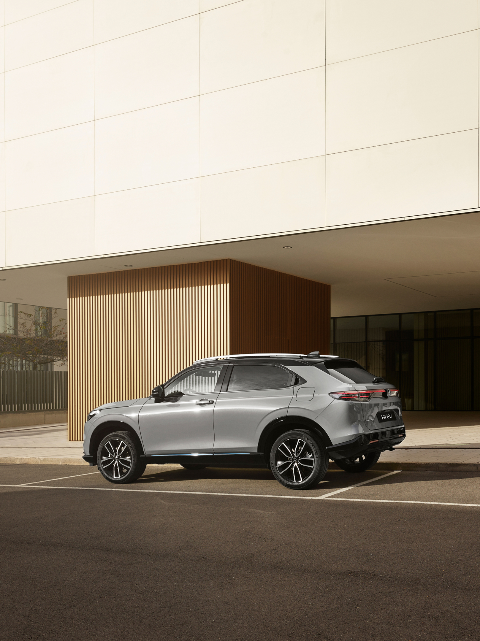 A silver Honda HR-V is parked in an outdoor parking space in front of a modern building with glass walls and vertical wooden paneling, photographed from a rear three-quarter angle in daylight.