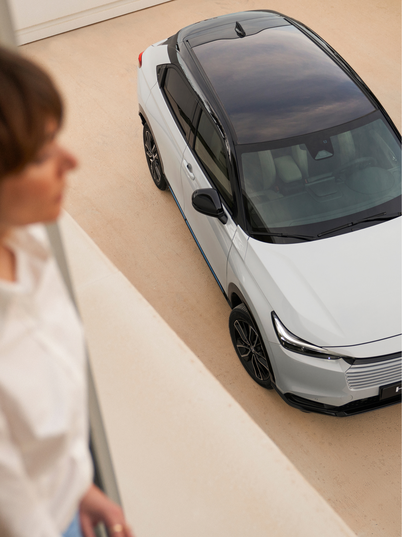 An overhead view of a white Honda HR-V with a black roof parked on a light-colored driveway, while a blurred person stands indoors in the foreground looking down toward the car.