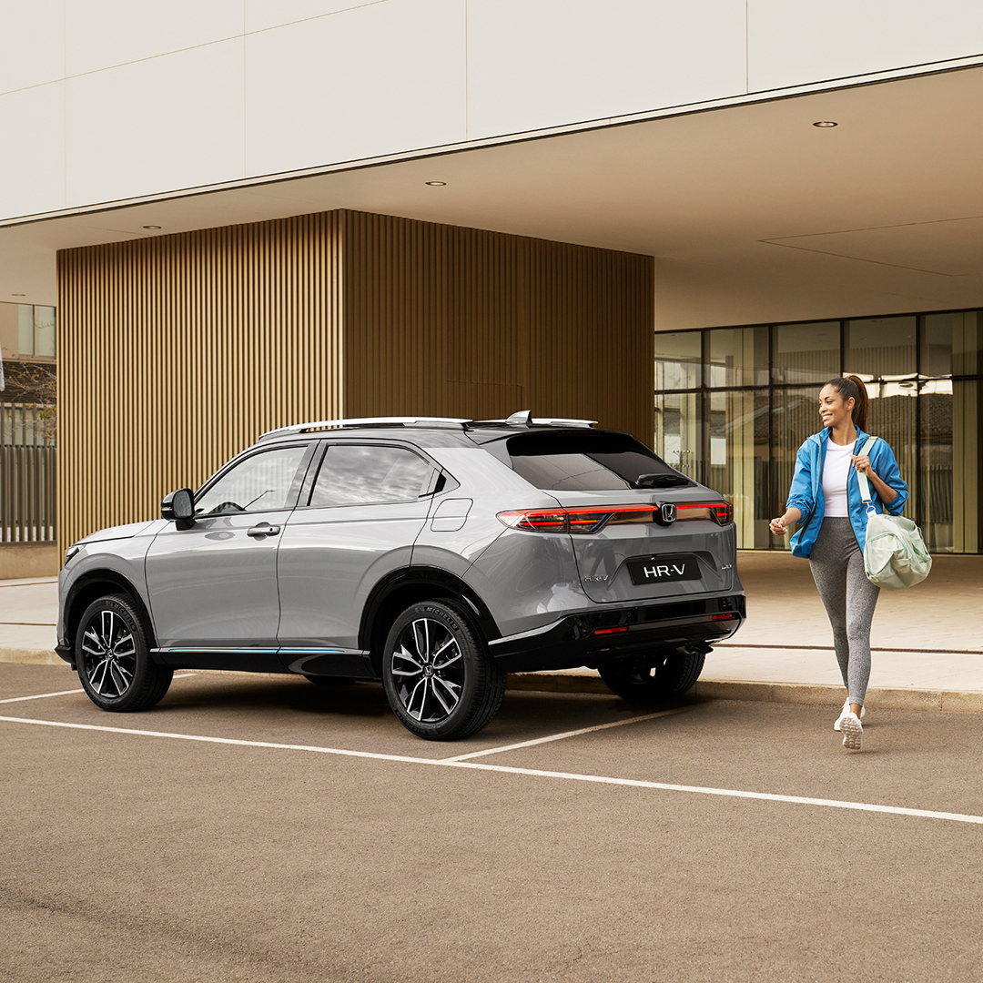 A silver Honda HR-V parked outside a modern building while a woman walks past it carrying a bag, suggesting everyday urban use and convenience.