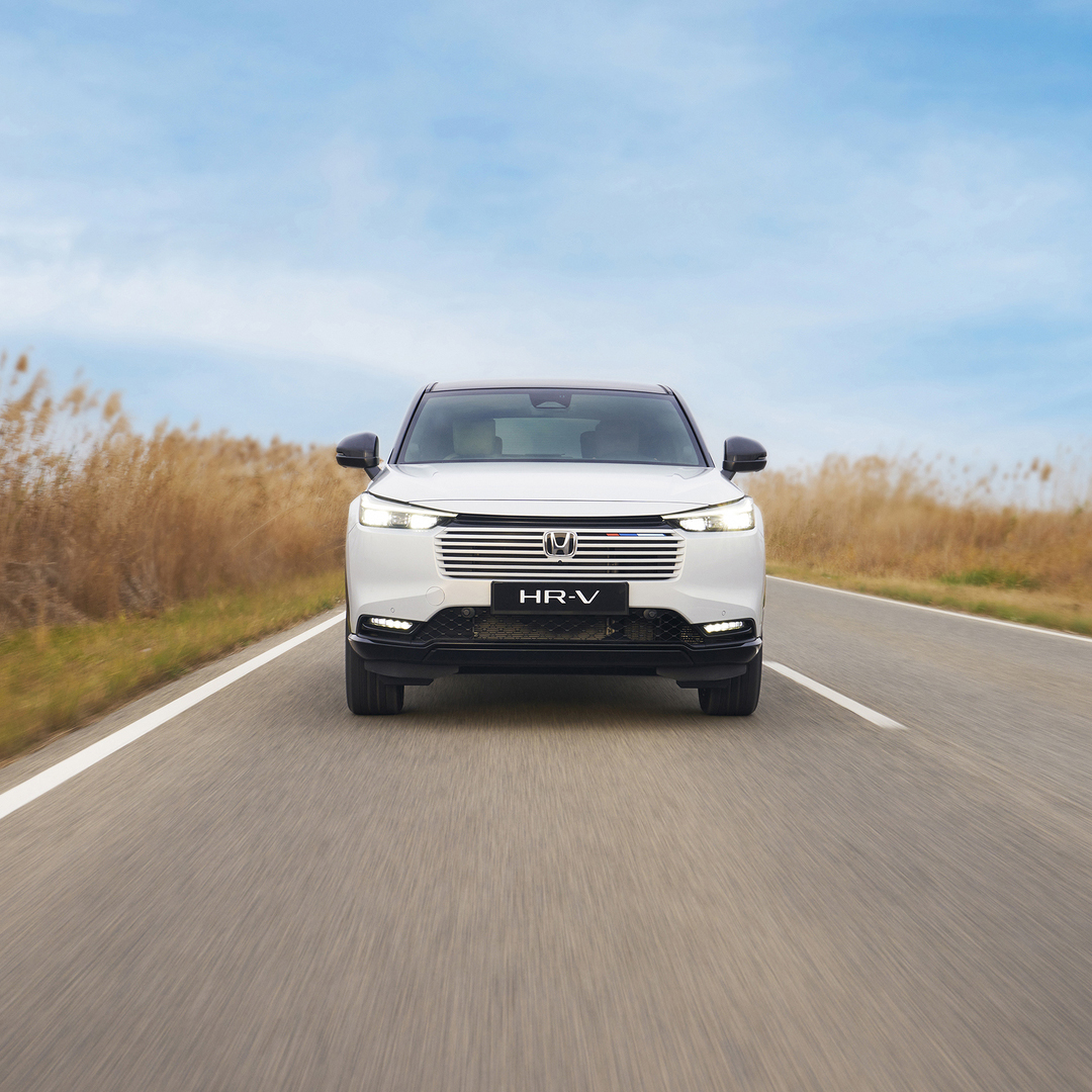 A white Honda HR-V drives toward the camera on a two-lane road, surrounded by tall golden grass under a blue sky.