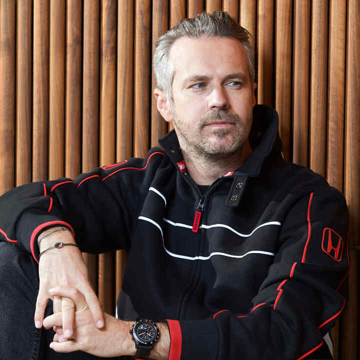 Man draagt Honda Racing pet en jas, raakt de rand van zijn pet aan.