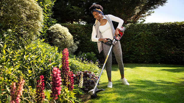 Vrouw gebruikt Honda accugazonstrimmer op gras in een tuin en demonstreert hoe de opvouwbare beschermkap planten beschermt.