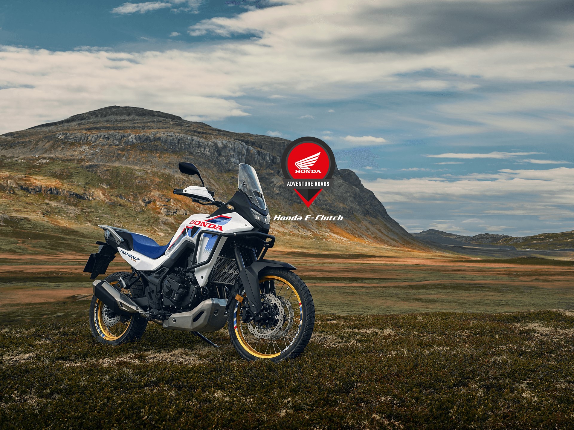 A Honda Africa Twin adventure motorcycle parked on rugged terrain with mountains in the background. The bike features a white, blue, and red color scheme. The Honda logo is superimposed in the top right corner of the image.