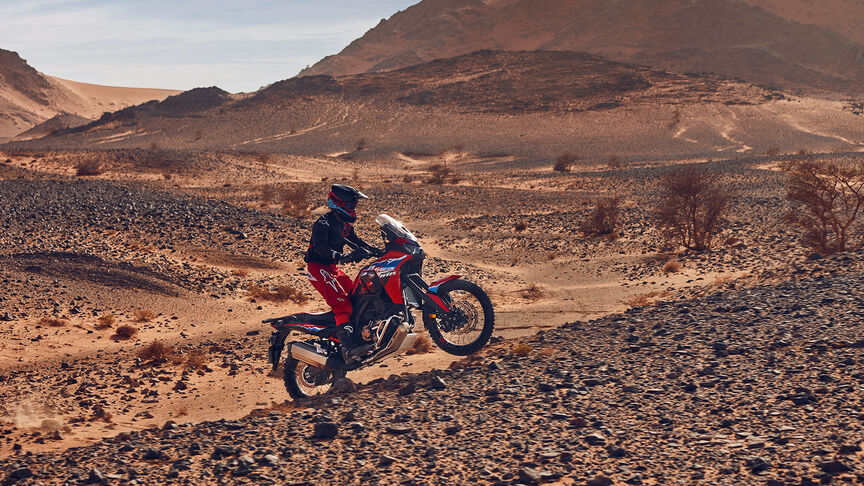 Model rijdend op een CRF1100L Africa Twin motorfiets op een woestijnlocatie.