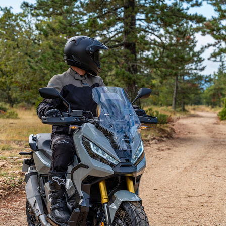 Honda X-ADV, driekwart vooraanzicht rechts, rijder staand op grijze motor, bosweg