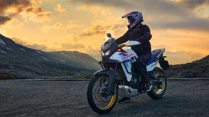 Model rijdend op een XL750 Transalp motorfiets op de weg in een berglandschap.