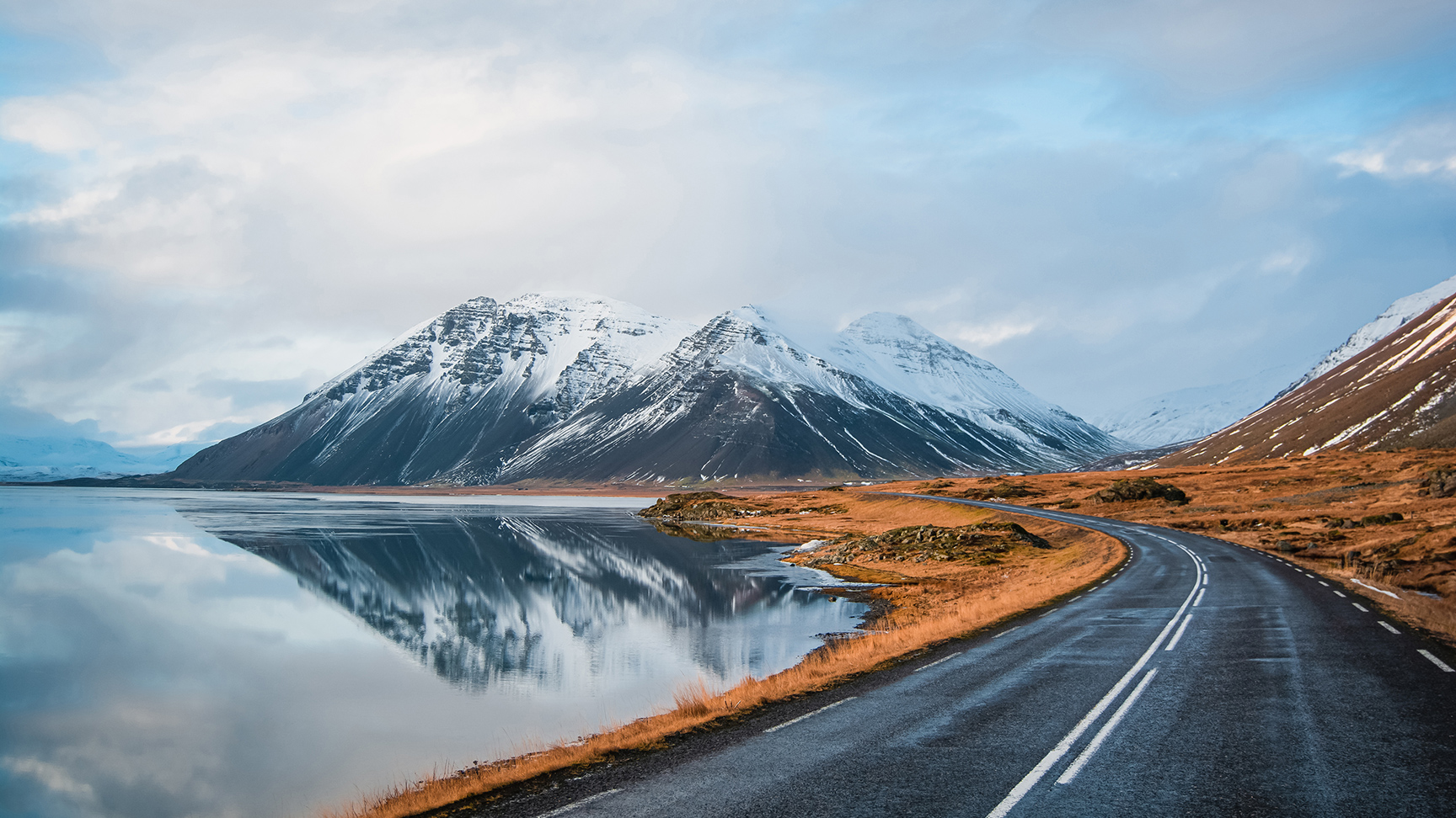 Panoramische winterfoto van weg die langs de kust van het meer naar vulkanisch gebergte leidt. Hoge, piekige rotsen bedekt met een sneeuwlaag die zich weerspiegelt in het wateroppervlak. Zo ziet de bestuurder de ringweg, IJsland.