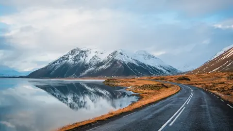 Panoramische winterfoto van weg die langs de kust van het meer naar vulkanisch gebergte leidt. Hoge, piekige rotsen bedekt met een sneeuwlaag die zich weerspiegelt in het wateroppervlak. Zo ziet de bestuurder de ringweg, IJsland.