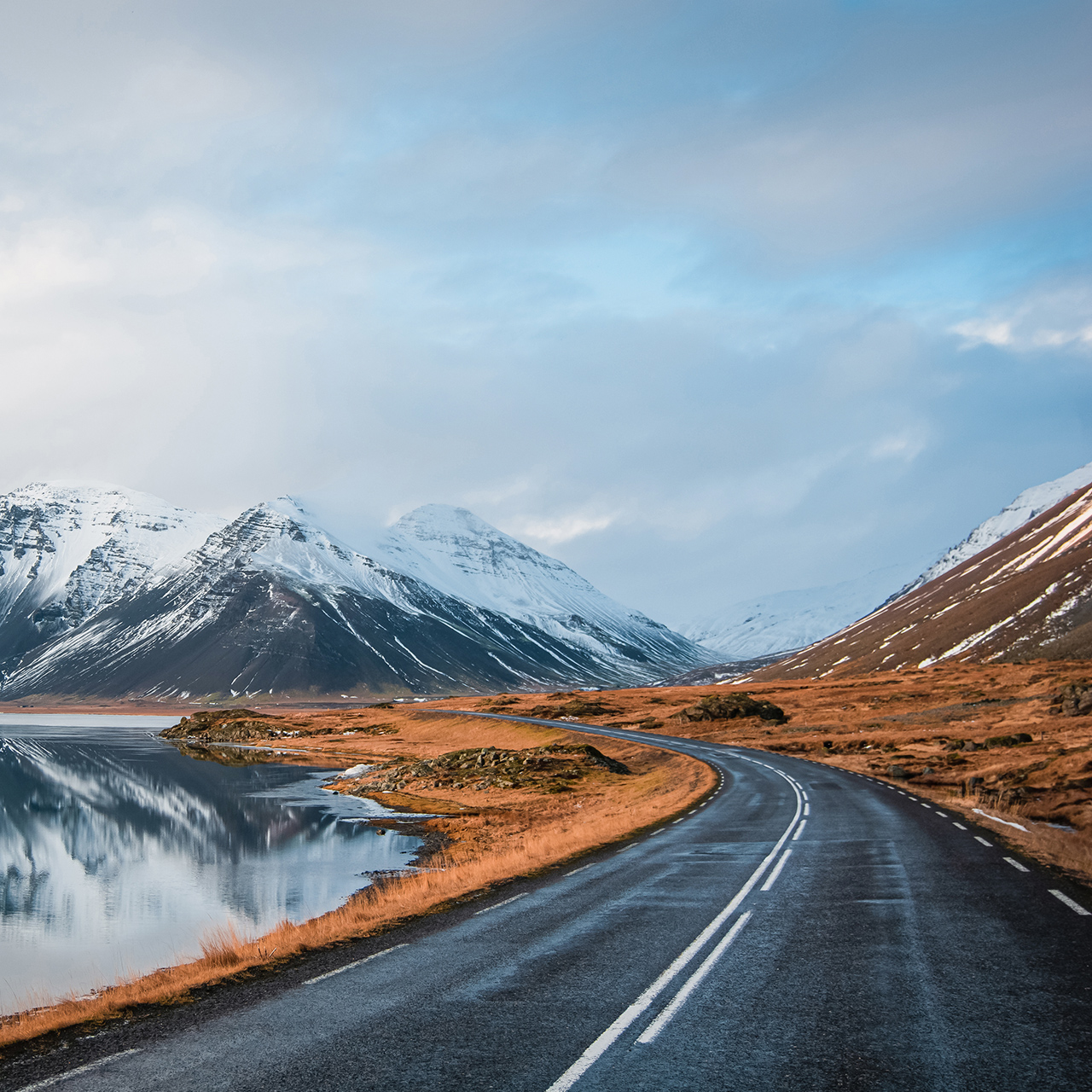 Panoramische winterfoto van weg die langs de kust van het meer naar vulkanisch gebergte leidt. Hoge, piekige rotsen bedekt met een sneeuwlaag die zich weerspiegelt in het wateroppervlak. Zo ziet de bestuurder de ringweg, IJsland.