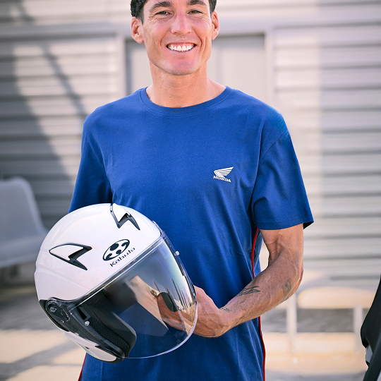 A smiling person wearing a blue Honda t-shirt holds a white motorcycle helmet. The Honda wing logo is visible on the shirt.