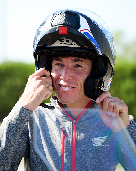 A person wearing a Honda-branded gray jacket adjusts a Kabuto motorcycle helmet. The helmet is predominantly white with blue and red accents. The individual is smiling while fastening the helmet strap.