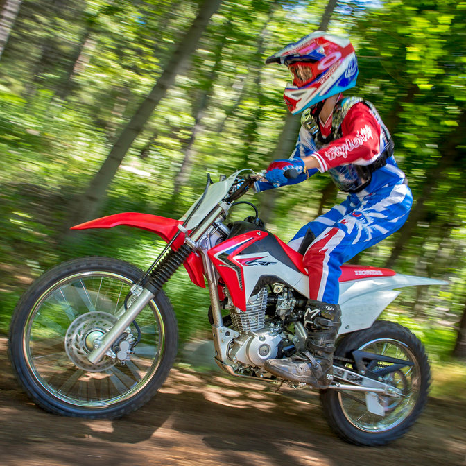 Zijaanzicht van CRF125F met rijder op onverharde weg op racelocatie in de bossen. Zicht op linkerkant.